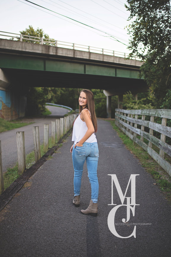 Woman standing on a paved path near a bridge wearing a white top and jeans