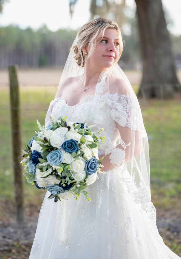 Bride in a white wedding dress holding a blue and white bouquet outdoors.