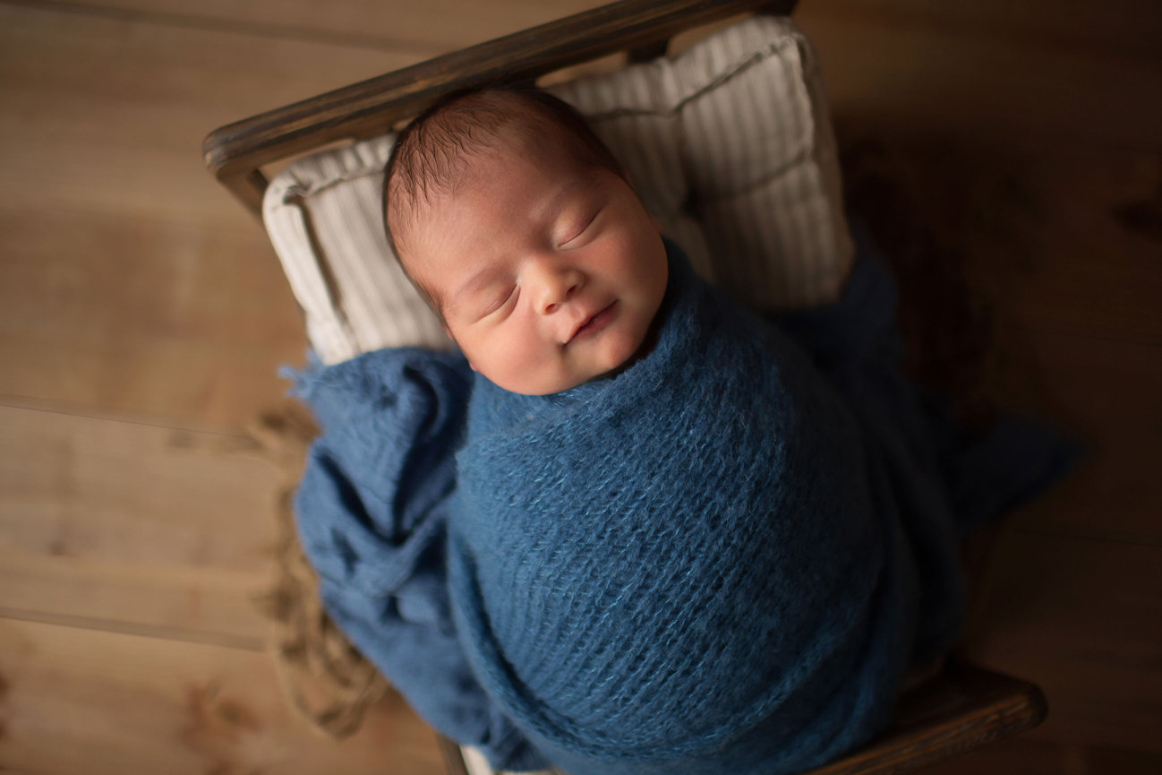 Newborn baby smiling in his sleep while wrapped in knit blue wrap and laying in tiny newborn bed on wooden backdrop in Kansas City Potography studio with Dreamworks Photography