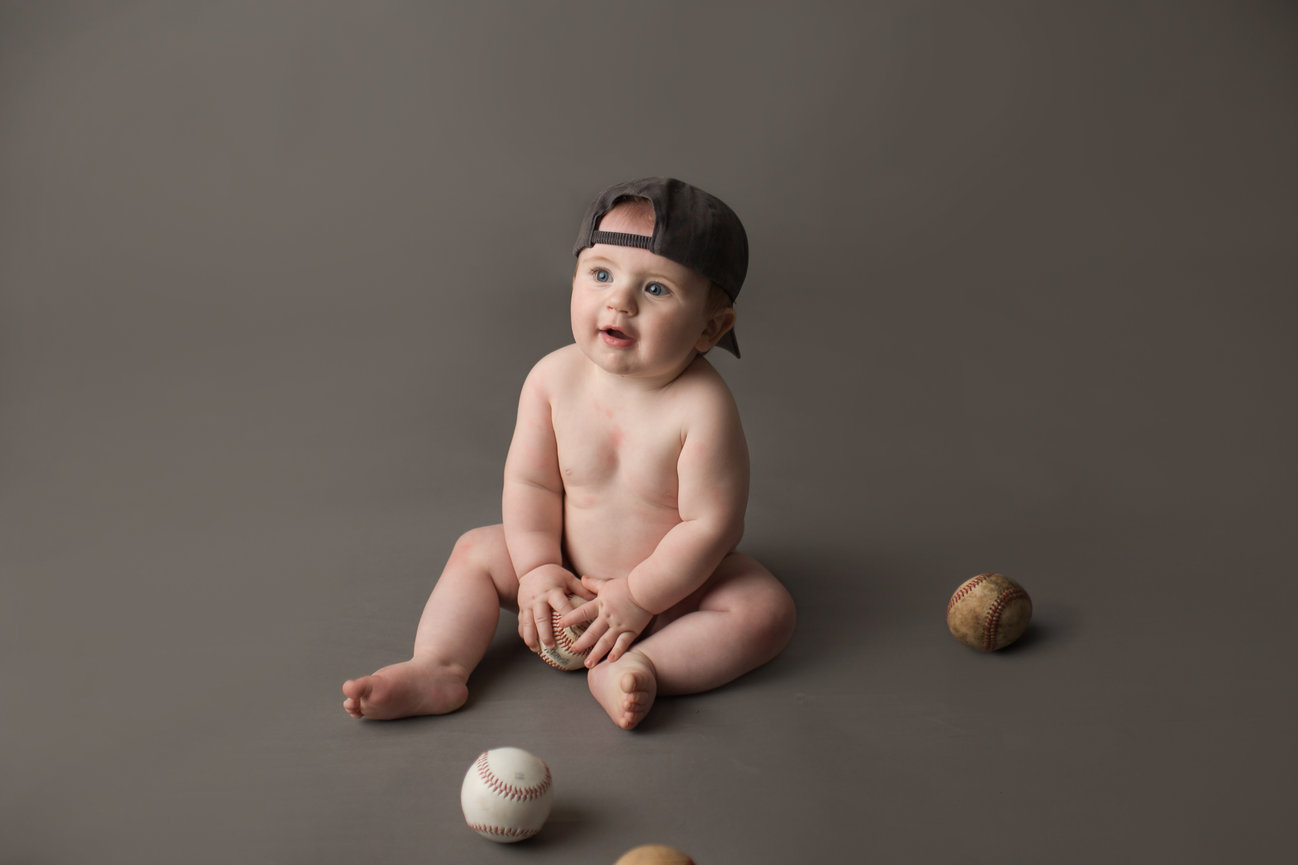 Baby boy with big blue eyes photographed on grey background wearing Kansas City royals hat holding baseball during milestone photo session with Kansas City baby photographer Dreamworks Photography