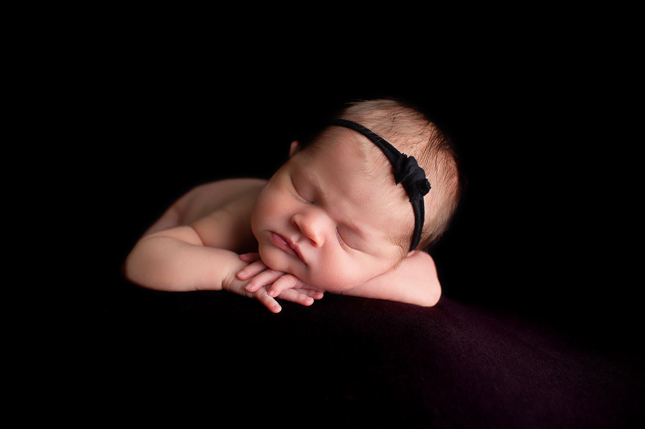 newborn baby girl sleeping in forward facing pose laying on her belly and chin resting on her hands with black background and tiny black headband with a bow during newborn photoshoot with newborn photographer Dreamworks Photography in Kansas City 