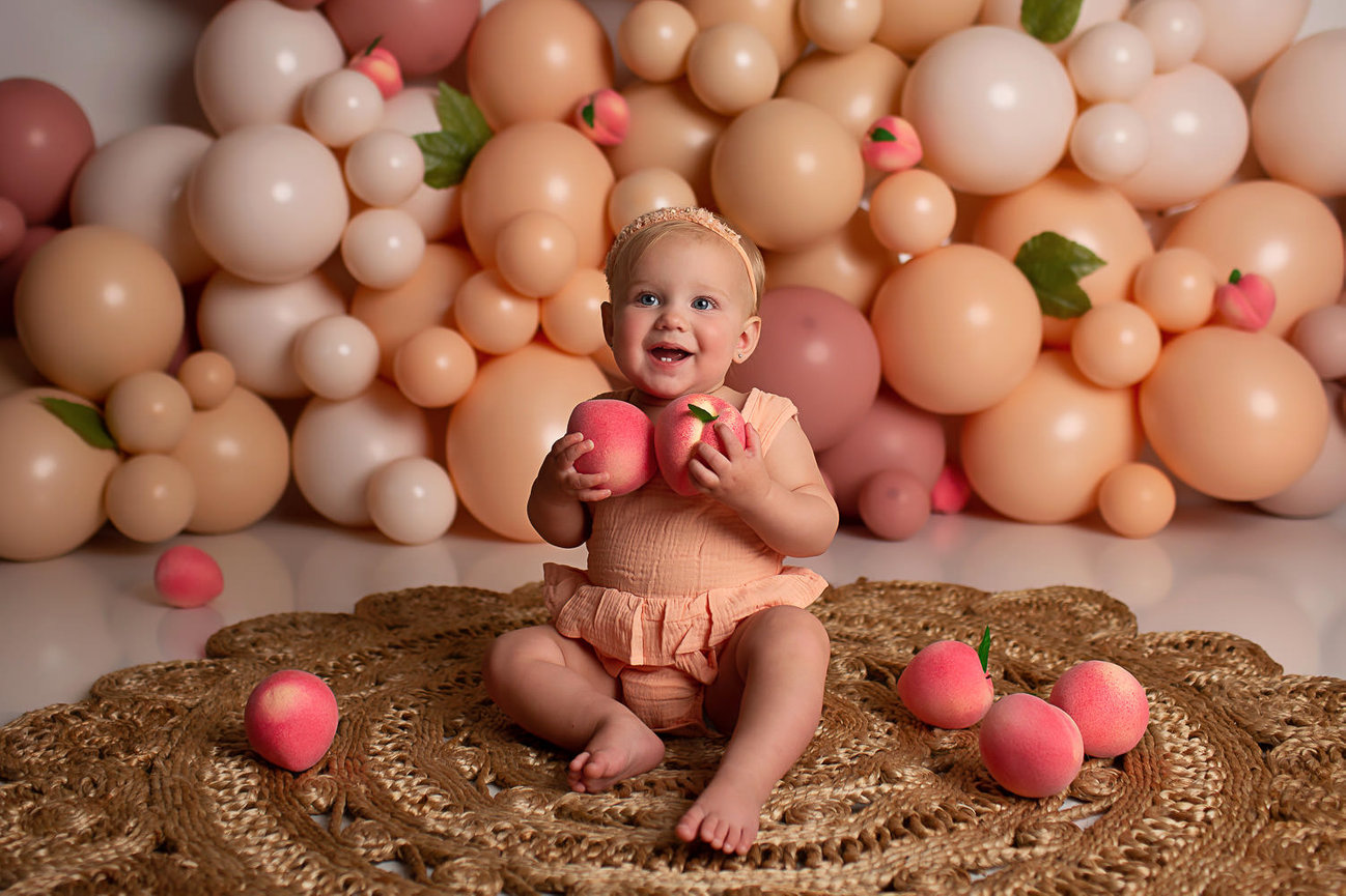 Baby girl sitting on boho just rug with peach pink and neutral colored balloons in the background during her first birthday cake smash photo session with Kansas City photographer Dreamworks Photography 