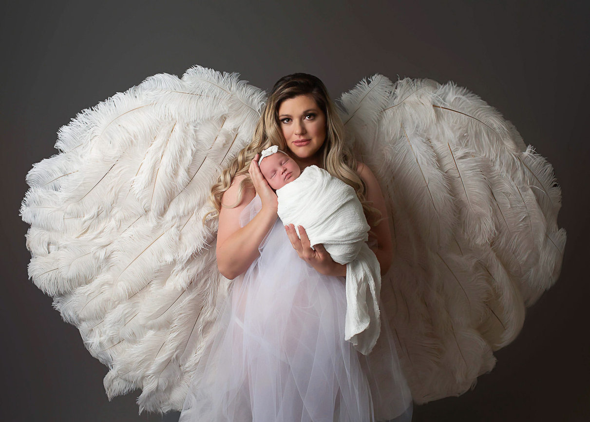 mother wearing white angel wings while holding newborn baby girl during newborn photoshoot with newborn photographer Dreamworks Photography in kansas city