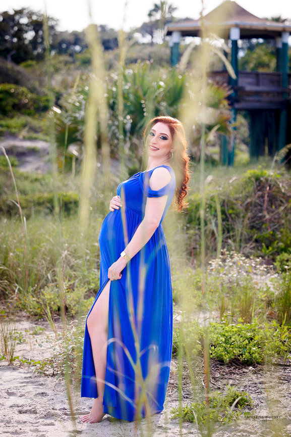 Pregnant woman in a blue dress stands barefoot on a sandy path with greenery and a wooden structure in the background during a maternity photography session with Catherine Whitney Photography in Jacksonville Florida