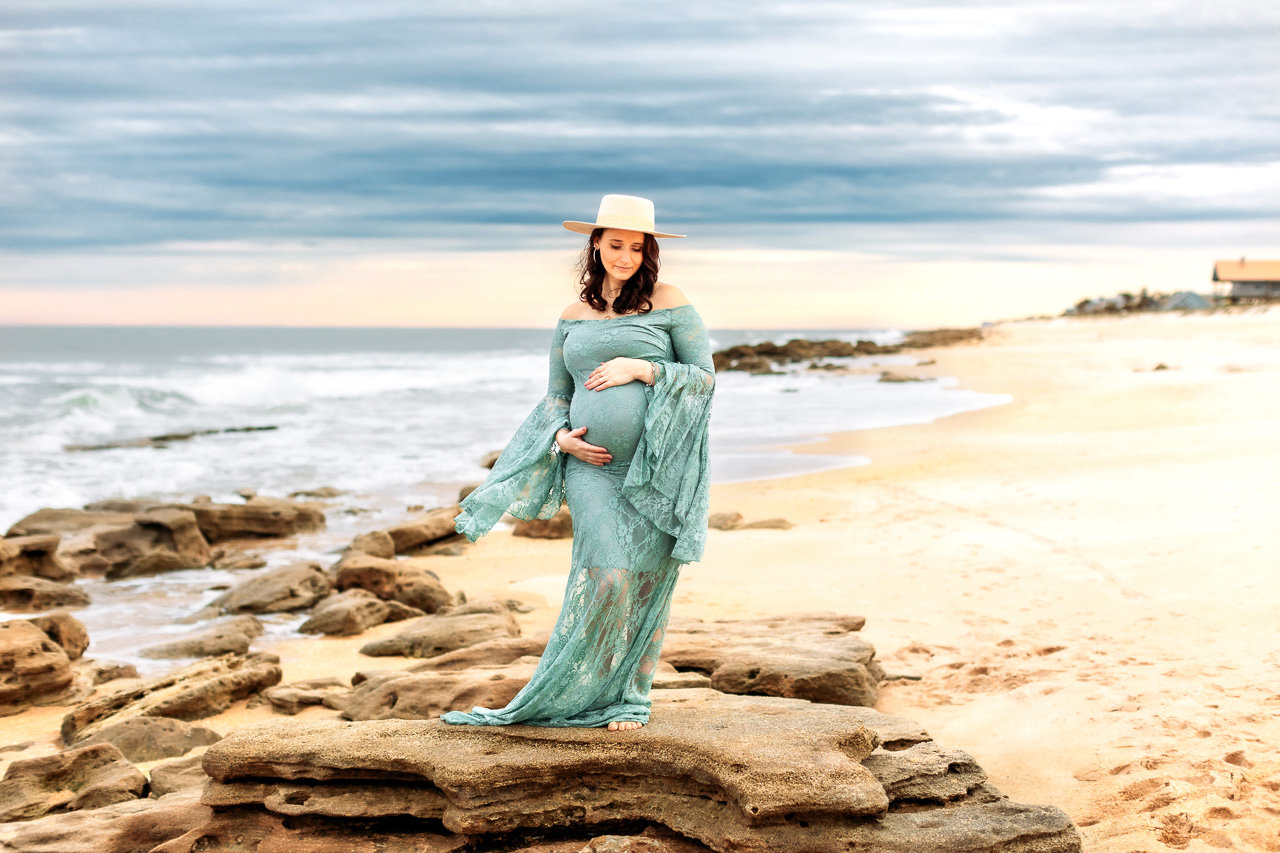 Woman in a flowy dress stands on rocky beach, holding belly and wearing a hat, with cloudy sky and ocean in background during a maternity photography session with Catherine Whitney Photography in St. Augustine Florida
