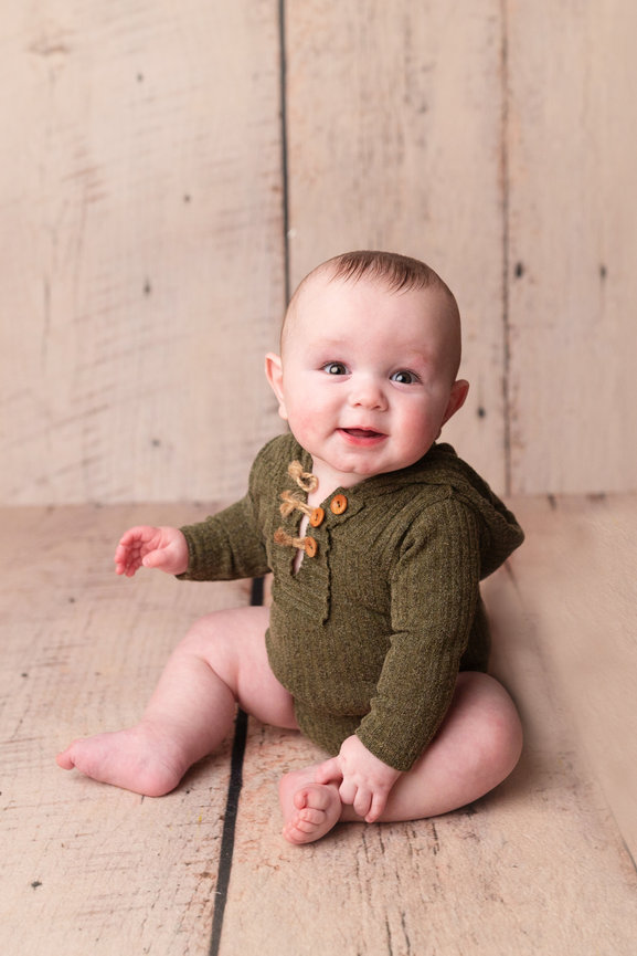 Omaha Baby Photographer photographs baby sitting on wooden floor, wearing a green knitted outfit with wooden buttons, smiling at the camera.