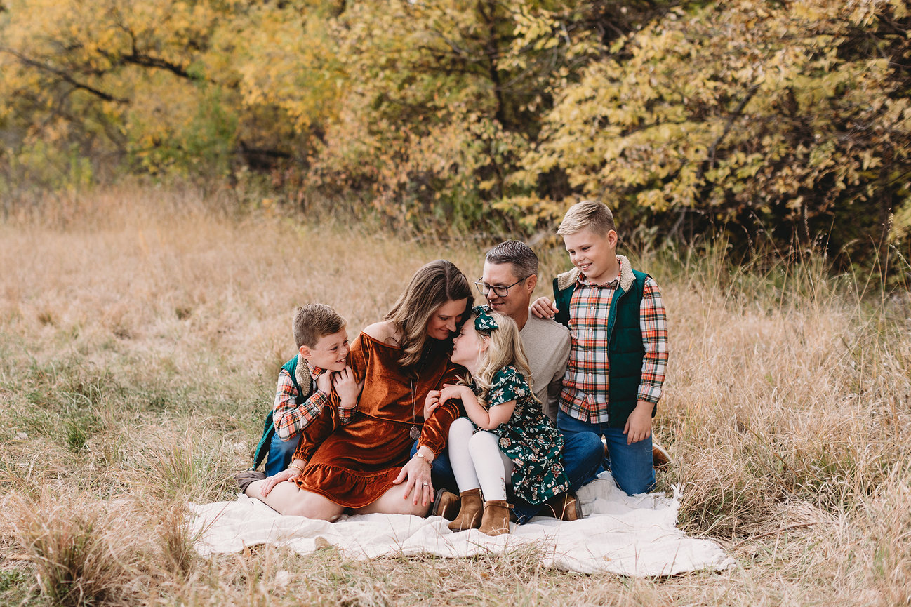 Family sitting on a blanket in a grassy field with autumn trees, wearing coordinated fall outfits, smiling and embracing.