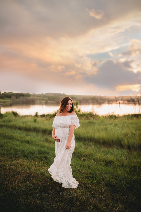 Pregnant woman in a white dress stands on grass by a lake at sunset with a colorful sky.