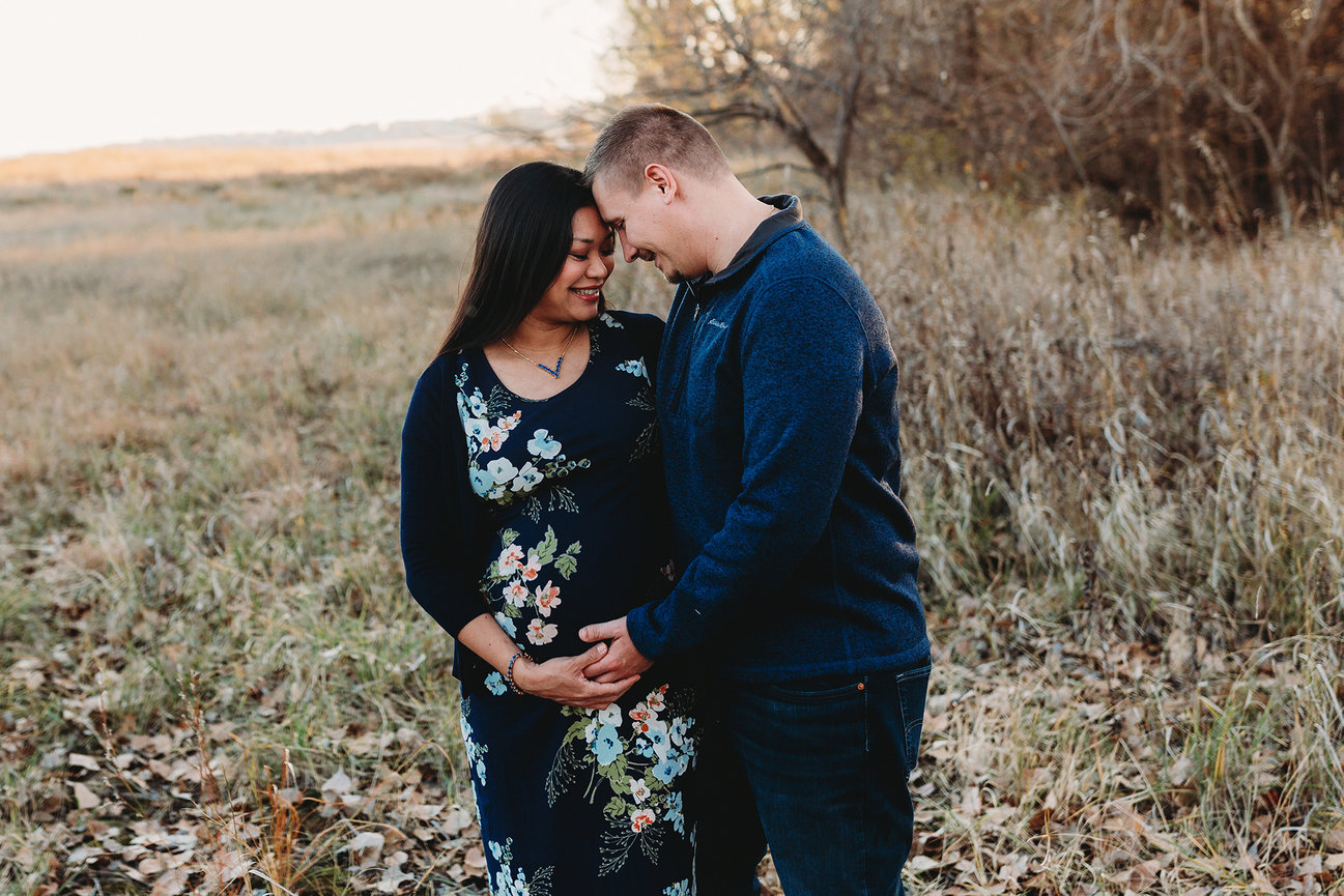 A couple in a field, with the man touching the woman's pregnant belly. They are smiling and standing close together.