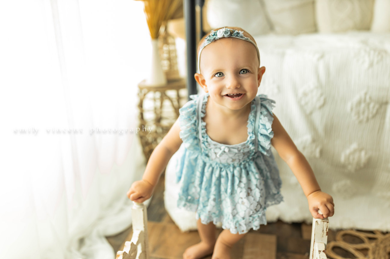 smiling baby girl with blue dress and headband posing in neutral bedroom setting photo session