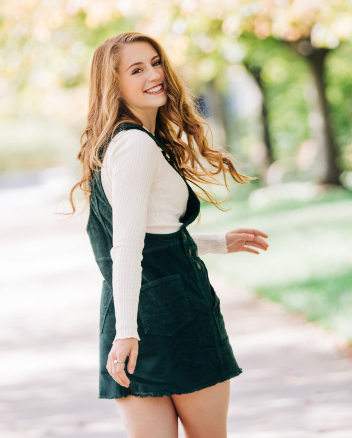 A high school senior woman with long hair in a white sweater and dark skirt strolling outdoors on a sunny day, with trees gently swaying in the background, conveying a sense of carefree graduation moments.