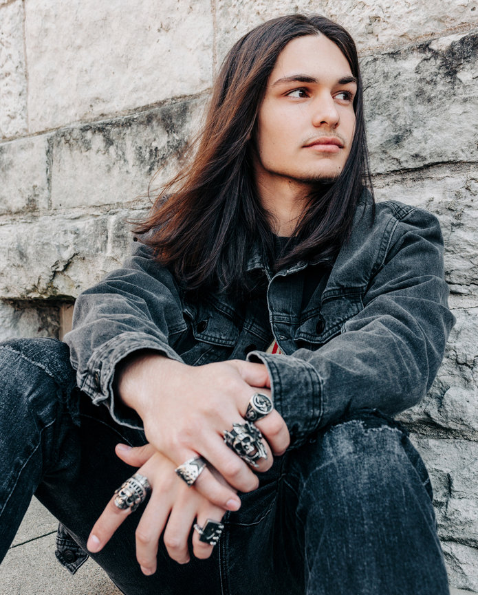 A high school senior with long hair and multiple rings sits against a stone wall, looking thoughtfully to the side in a dark denim jacket.