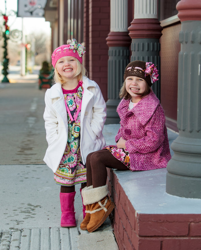 Two smiling children in colorful winter outfits sitting on a brick ledge outside, with a building and sidewalk in the background.
