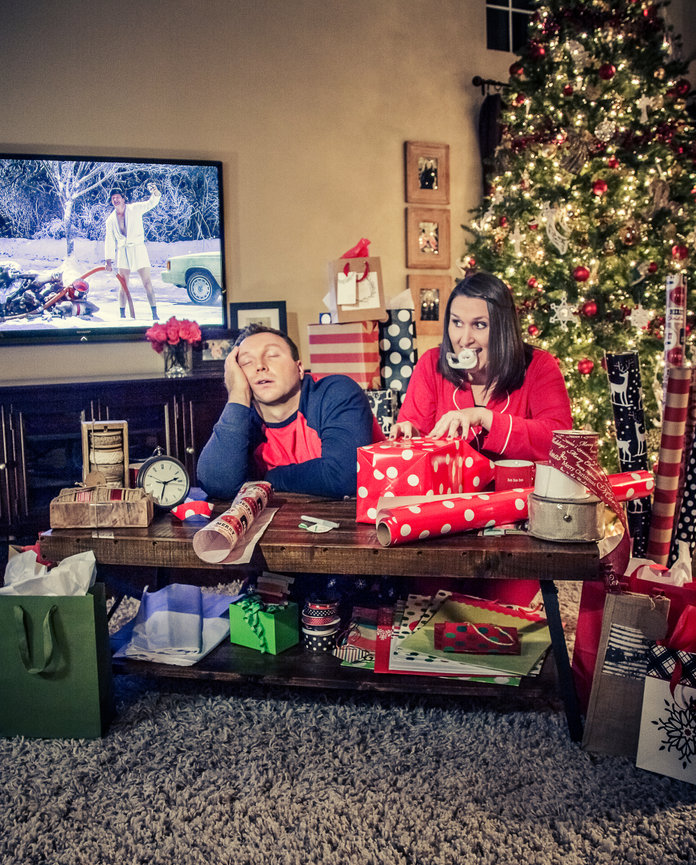 Couple sitting tiredly at a table with Christmas gifts and wrapping paper; Christmas tree and TV in the background.