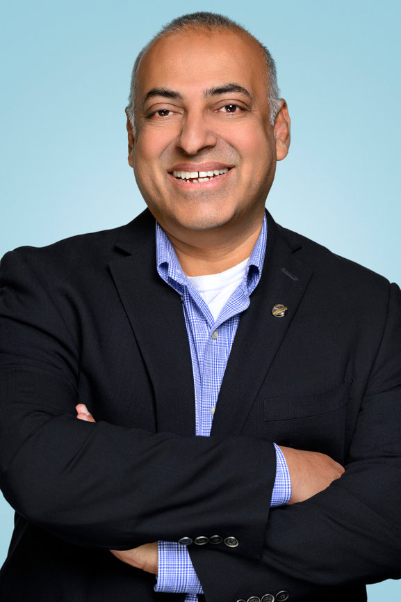 Smiling man in a black blazer with arms crossed, set against a light blue background.