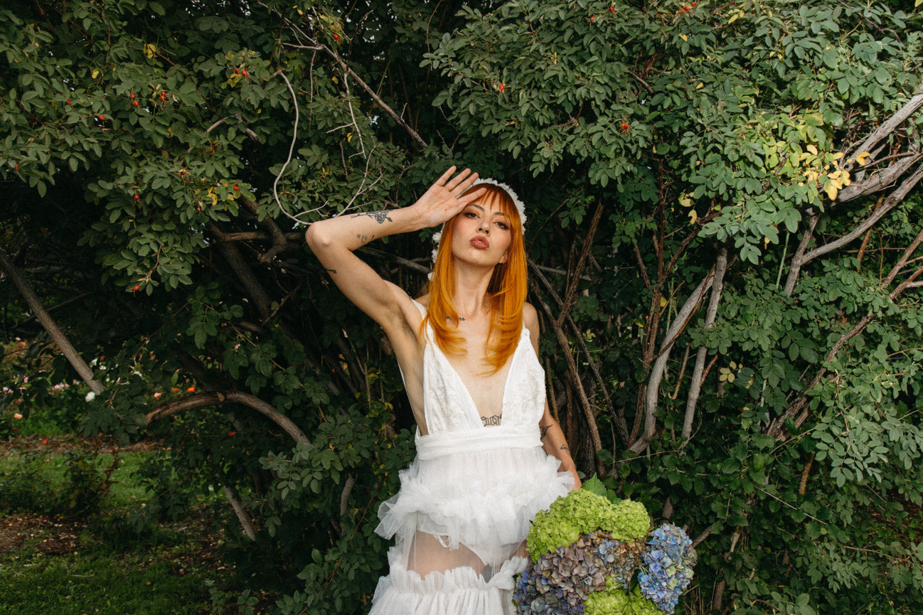 Person with orange hair in a white dress standing by a leafy tree, holding hydrangeas.