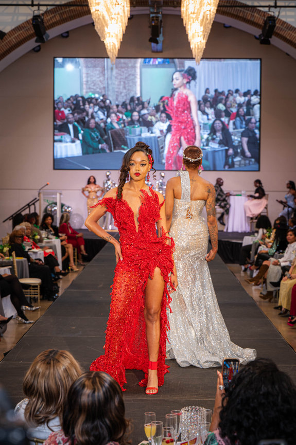 A fashion model in a red dress walks on a runway, with an audience and large screen in the background captured in the style of Jo Hayes Images Charlotte Event Photography Charlotte Portrait Fashion Photographer on location The Revelry Camp North End