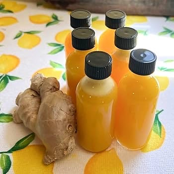 Six small bottles of orange liquid next to a ginger root on a lemon-patterned tablecloth.