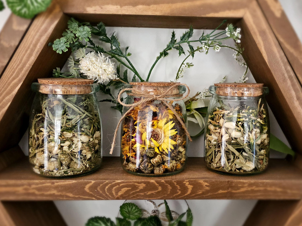 Three glass jars with cork lids filled with dried herbs and flowers, displayed on a wooden shelf with green plant accents.