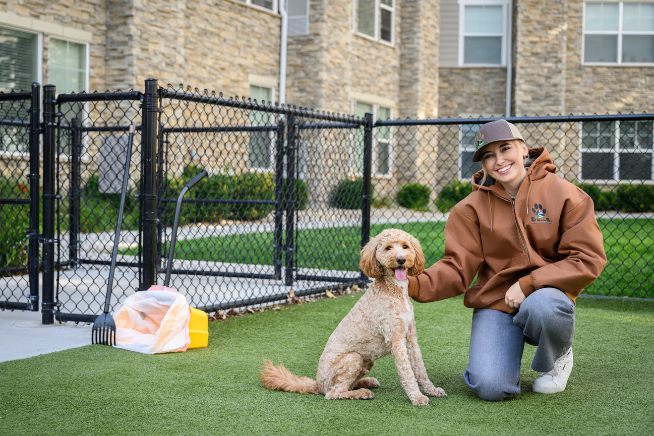 Person kneeling on grass beside a happy dog with a chain-link fence and brick building in the background.