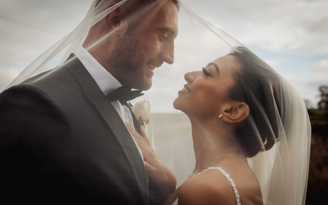 Bride and groom smiling under a veil during their wedding portrait