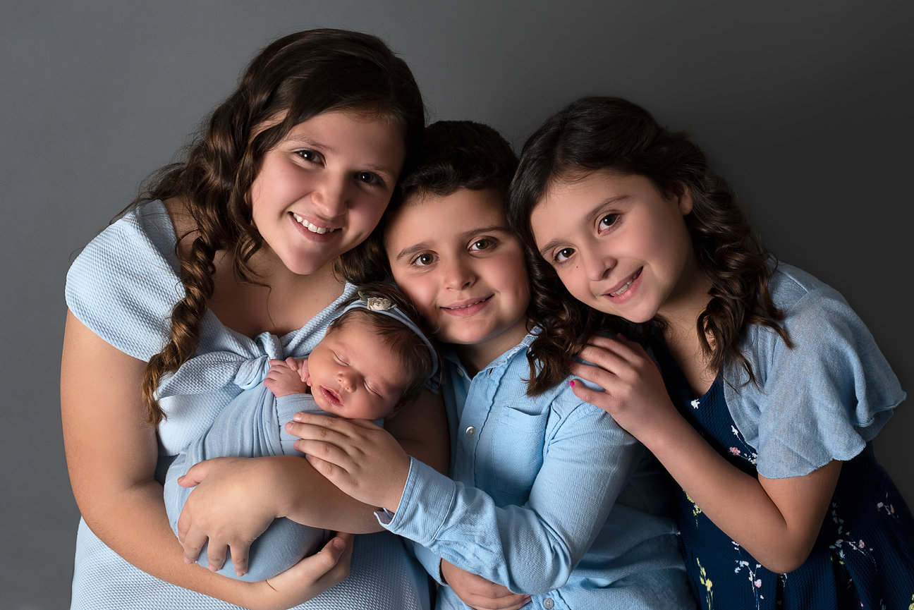 Four children pose together against a gray background. Three older kids, two girls and one boy from Tower Lakes, Illinois, smile while the older girl gently holds a sleeping baby. All are dressed in shades of blue and have wavy, dark hair.