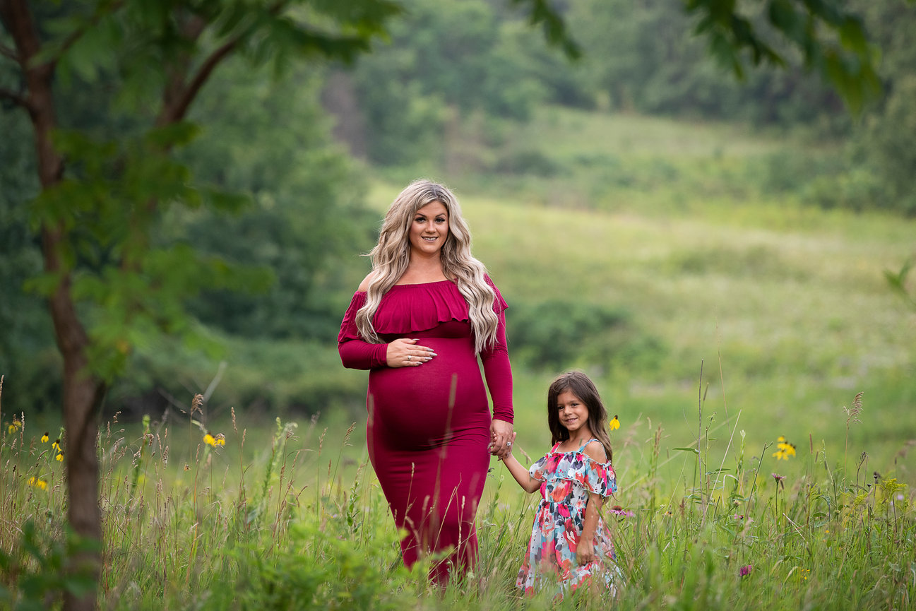A pregnant woman in a red dress holds hands with a young girl in a floral dress. They are standing in a lush green field surrounded by wildflowers, under a canopy of trees. The scene conveys a sense of warmth and family connection.