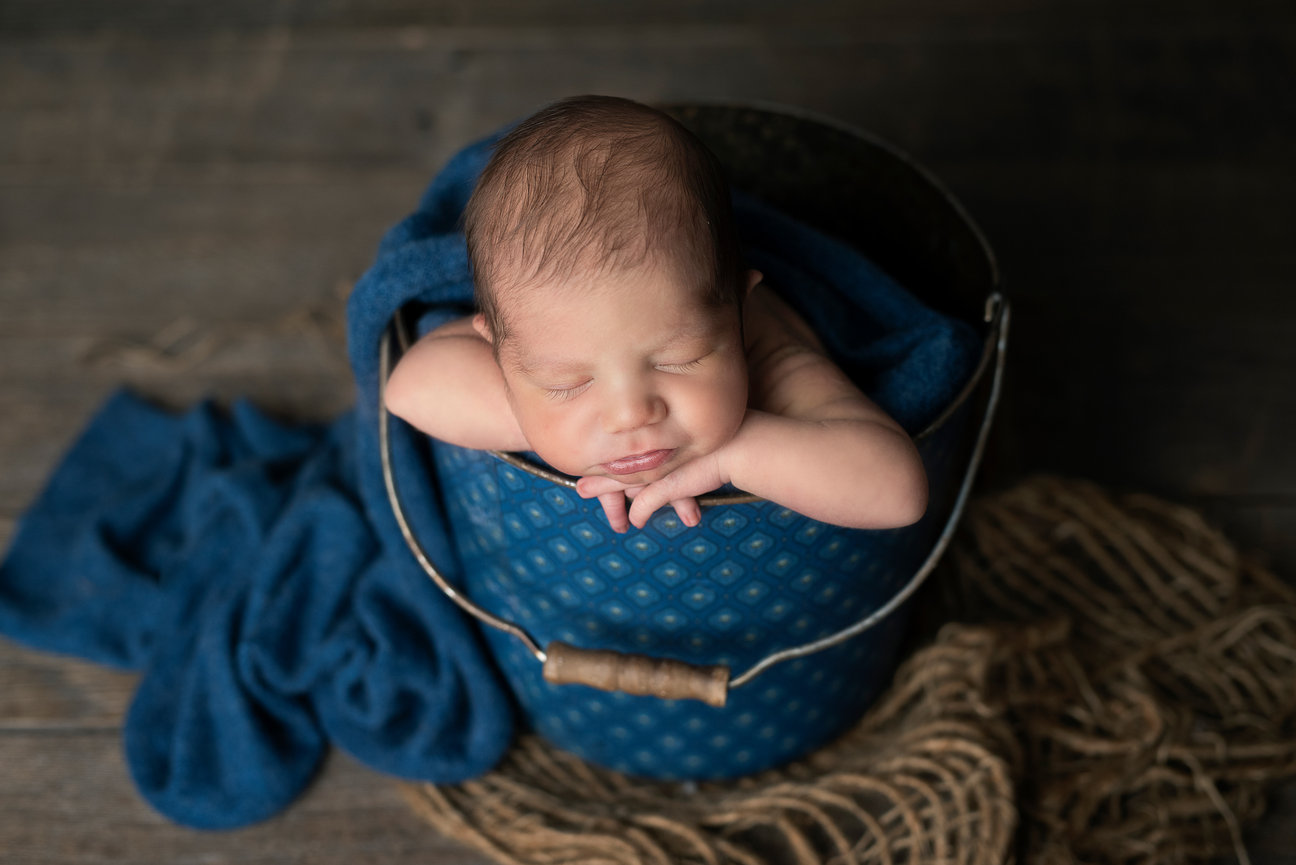 A sleeping newborn rests in a blue patterned bucket with a handle, their head and arms draped over the edge. The scene, set on a textured surface with blue fabric nearby, evokes the tranquil charm one might find in Elk Grove Village, Illinois.