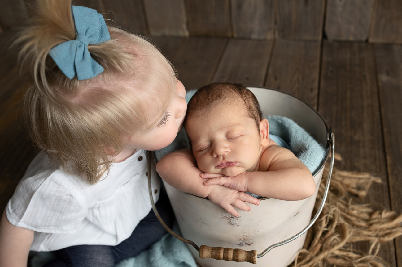 In a charming scene on a rustic wooden floor in Carol Stream, Illinois, a toddler with a blue bow gently kisses a sleeping baby nestled in a bucket with a cozy blue blanket.