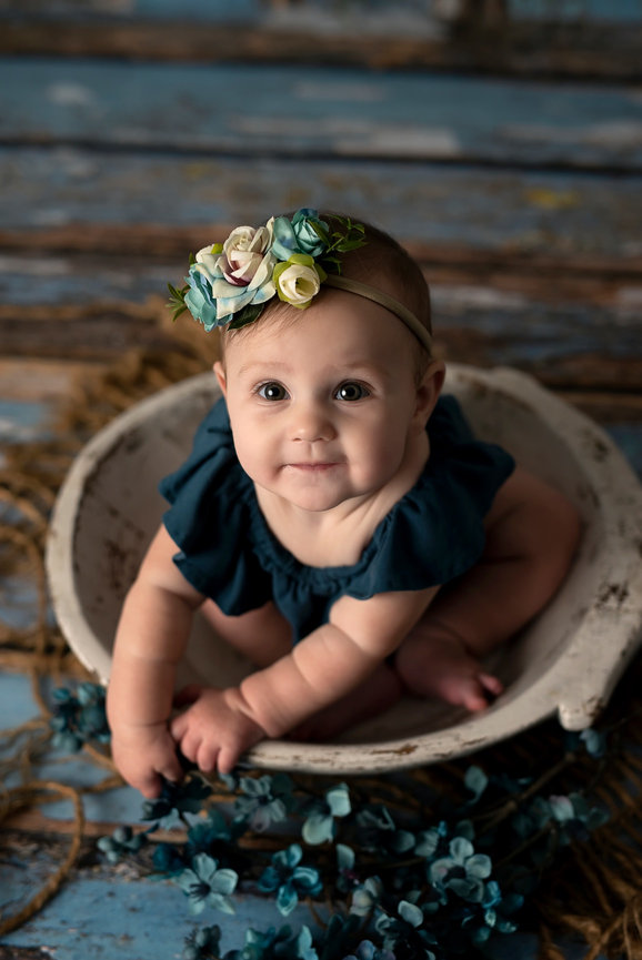 A nine-month-old baby, wearing a blue outfit and a floral headband, sits in a rustic white bowl surrounded by scattered blue flowers on a textured wooden floor. The baby is smiling and looking up.