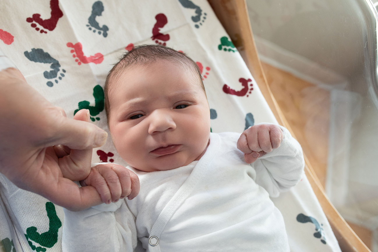 A newborn baby, swaddled in a white outfit, lies peacefully on a blanket adorned with colorful footprints. At Woodstock Hospital in Illinois, the infant gently grips an adults finger with both hands, displaying a serene expression.