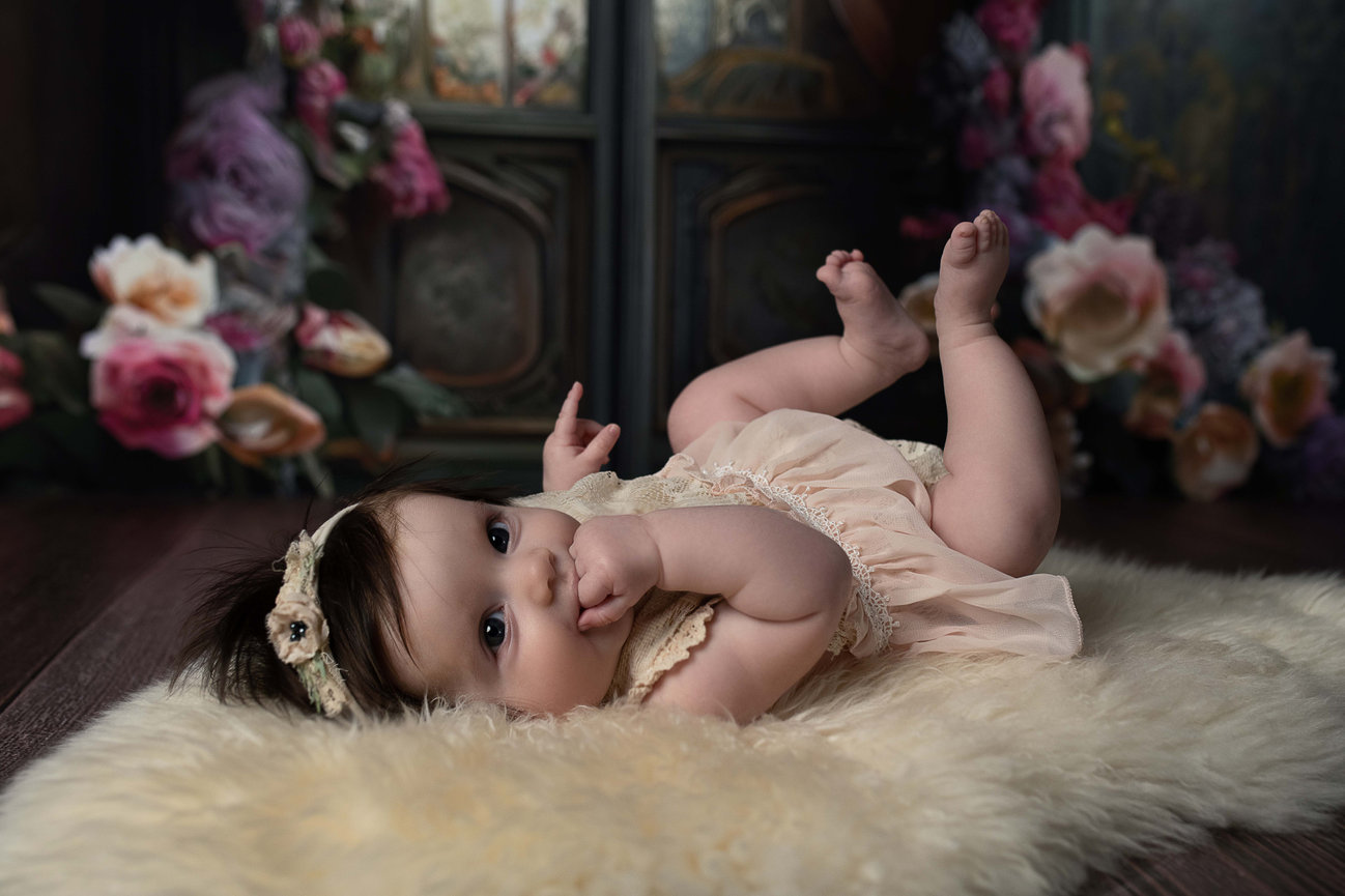 A baby photographer captures a moment where a baby, dressed in a light-colored romper and headband, lies on a fluffy blanket. With one hand near its mouth, the little one is surrounded by roses and other flowers against a softly lit backdrop.