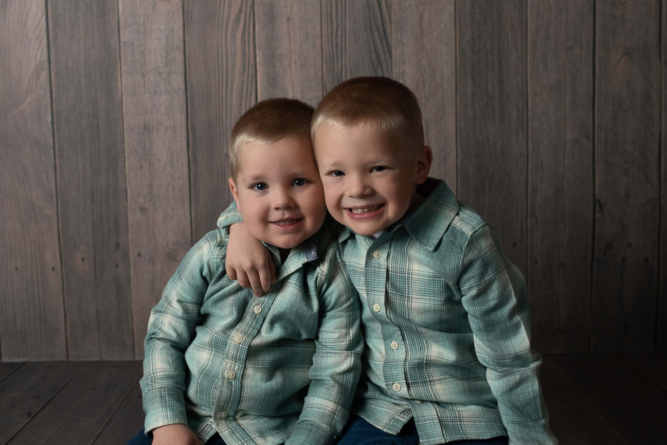 Two young boys with short hair sit close together, smiling warmly in a studio photography setting. They wear matching plaid shirts in shades of green and blue, and one boy has his arm around the others shoulder.