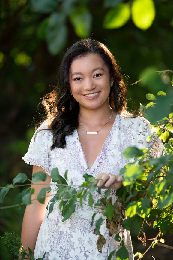 A teenager with long hair smiles gently while standing outdoors in Chicago. She is wearing a white lace dress and holding a green leafy branch. The background is softly blurred with lush greenery, suggesting a natural setting.