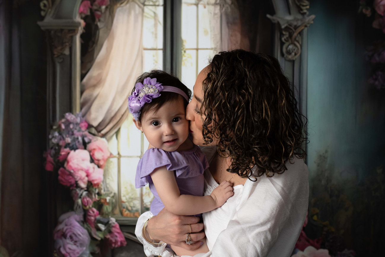 In a tender moment of child photography, a woman lovingly holds and kisses a baby. The little one, in a purple dress and headband, sits before an ornate window surrounded by colorful flowers, crafting a soft and warm atmosphere.
