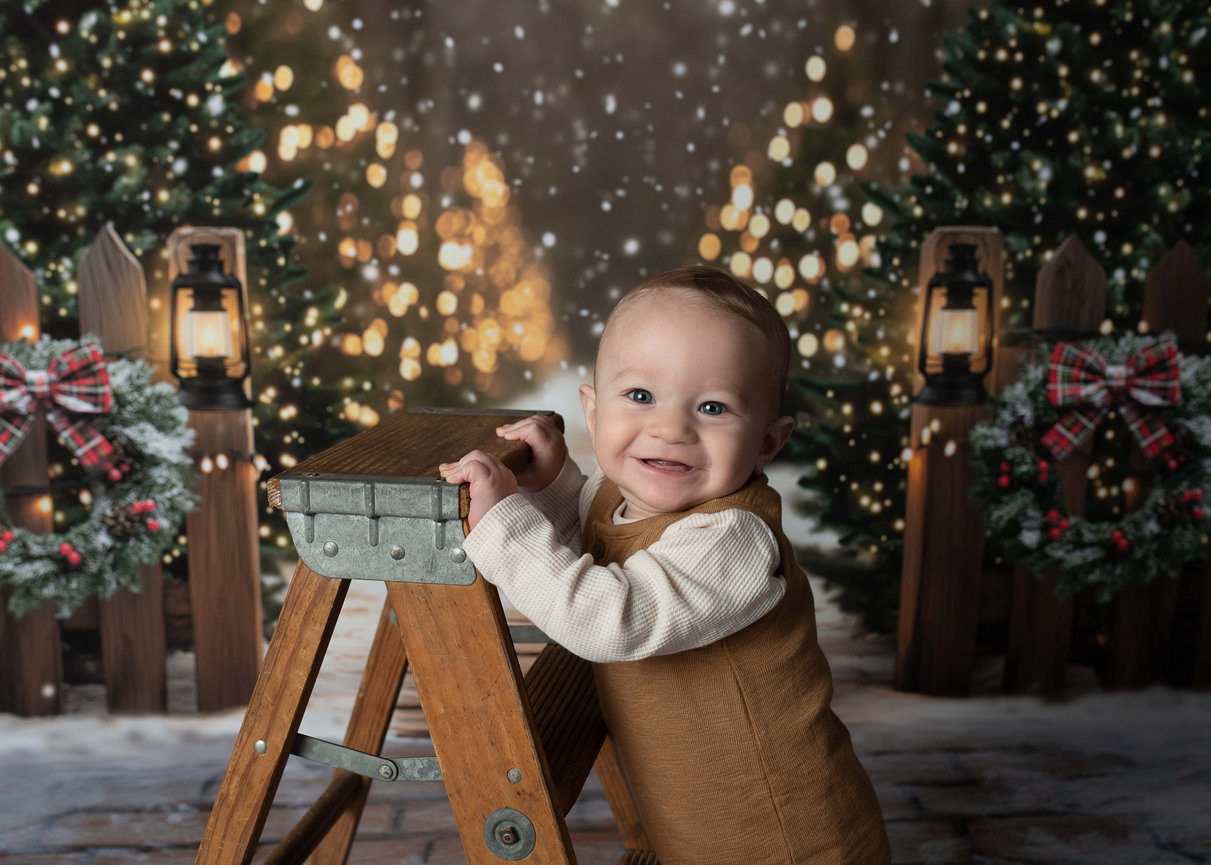 A smiling 9-month-old baby stands next to a wooden stepladder in front of a festive backdrop, capturing the essence of Christmas photography with twinkling trees and glowing lanterns.