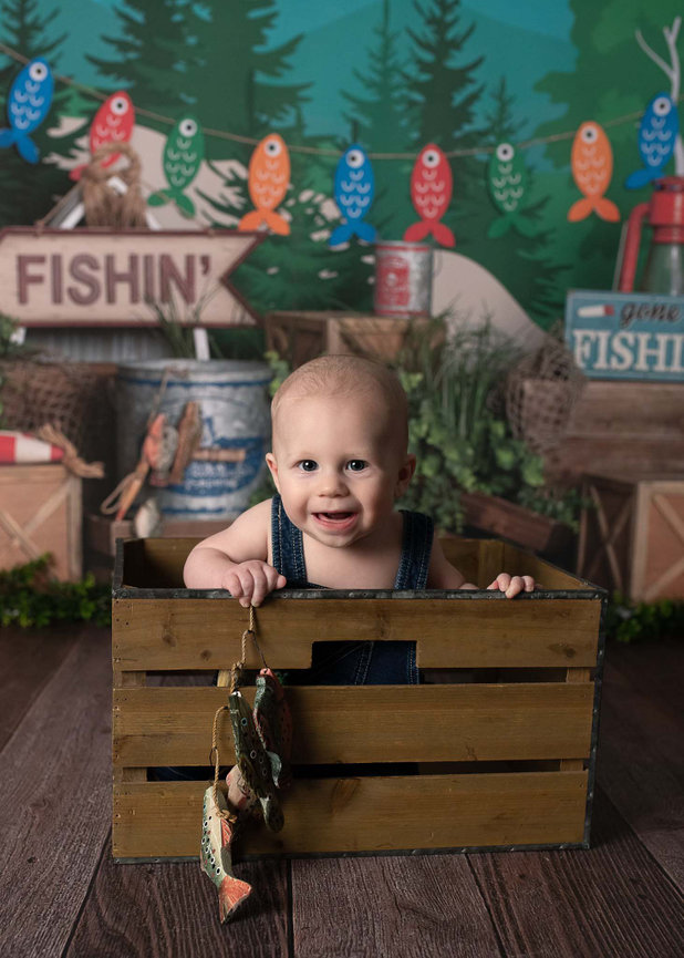 A 6-month-old baby in denim overalls smiles while standing in a wooden crate. The background has a fishing theme with props like a Gone Fishin sign, colorful fish decorations, and fishing gear.