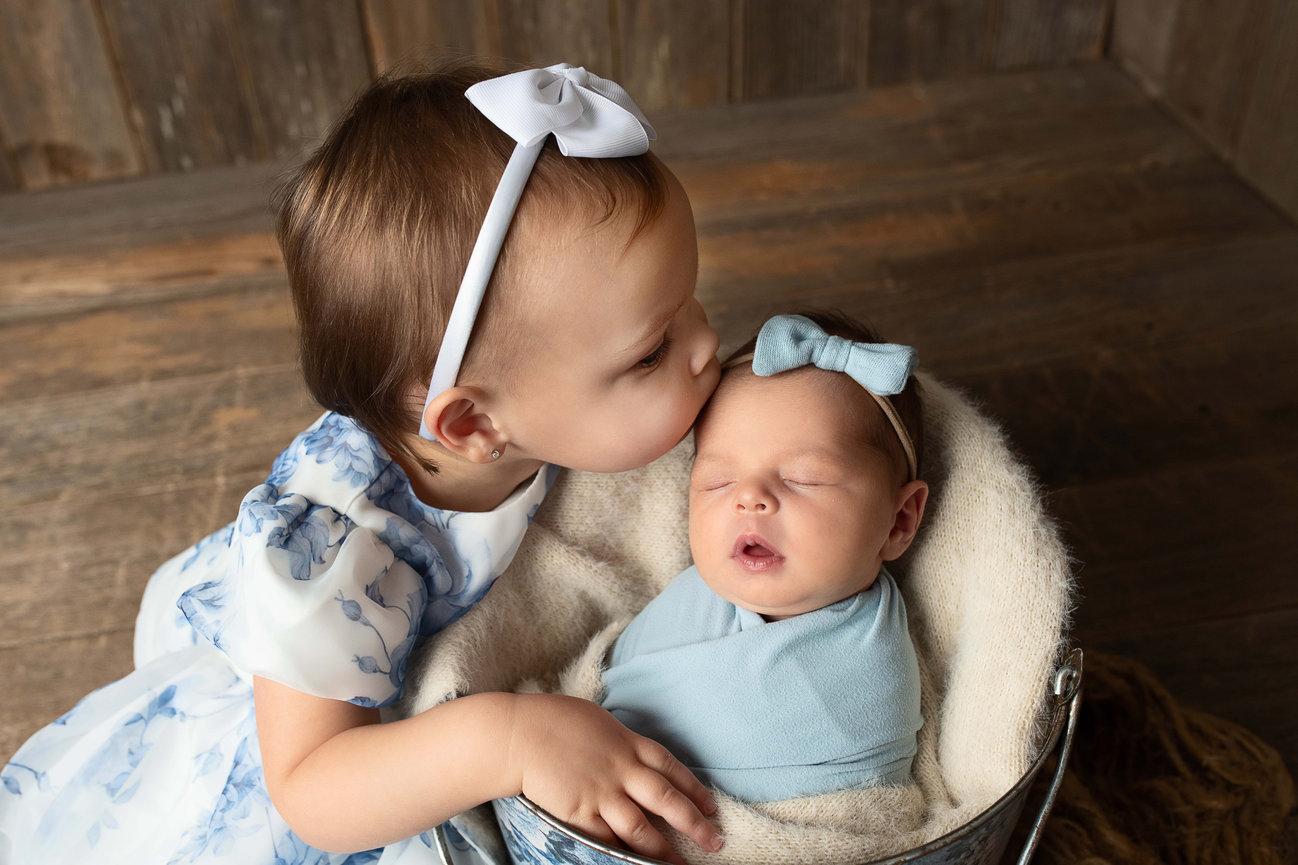 A young child with a white headband kisses a sleeping baby wrapped in a light blue blanket, adorned with a matching blue bow. Nestled in a cozy basket on the wooden floor, this tender moment captures the warmth of family life in Woodstock, Illinois