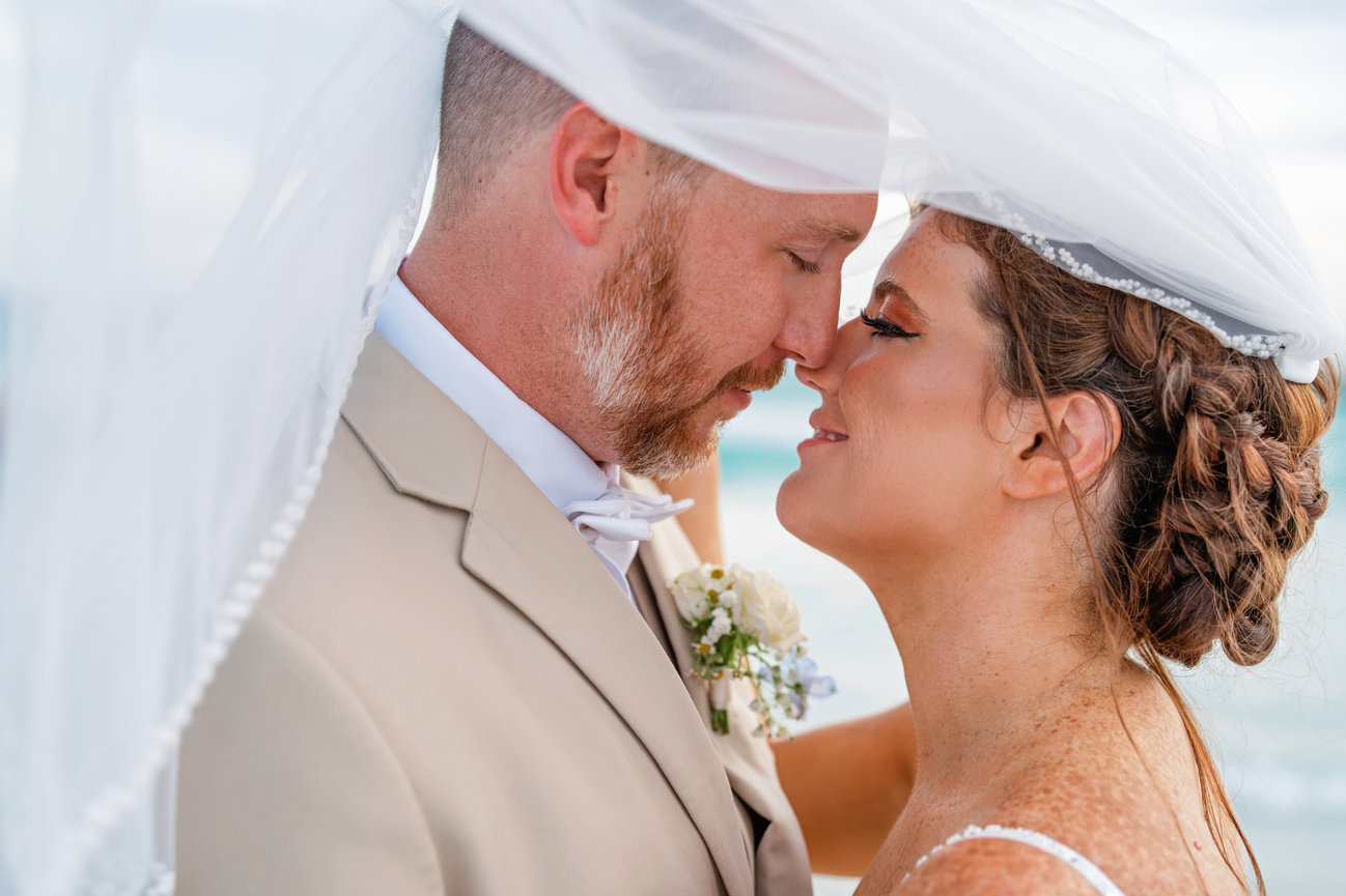 Bride and groom embracing under a veil with a beach background.