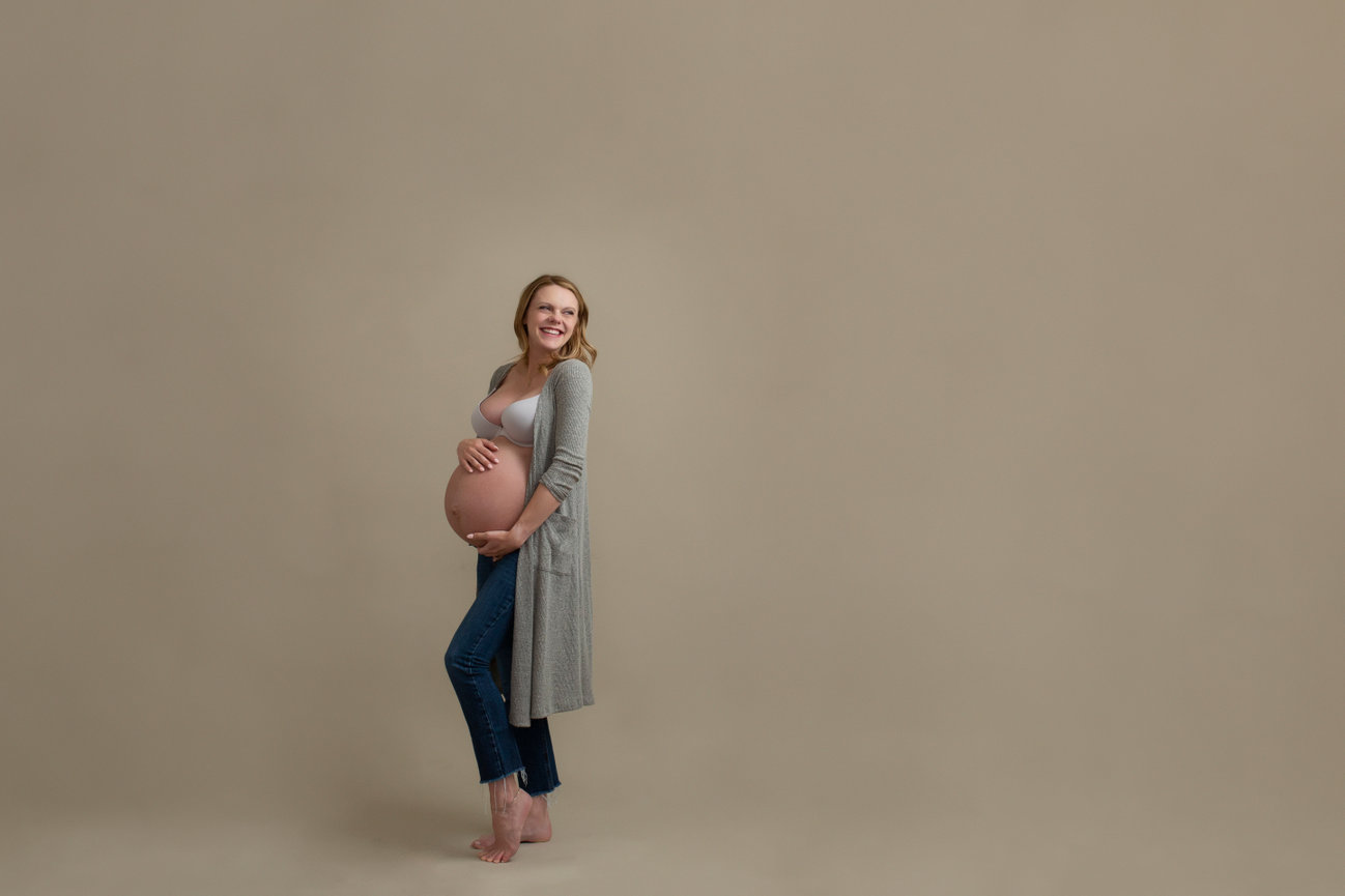 Fun studio maternity photo of woman holding her bare pregnant belly, looking up and back while laughing. She is wearing jeans, and a long cardigan.
