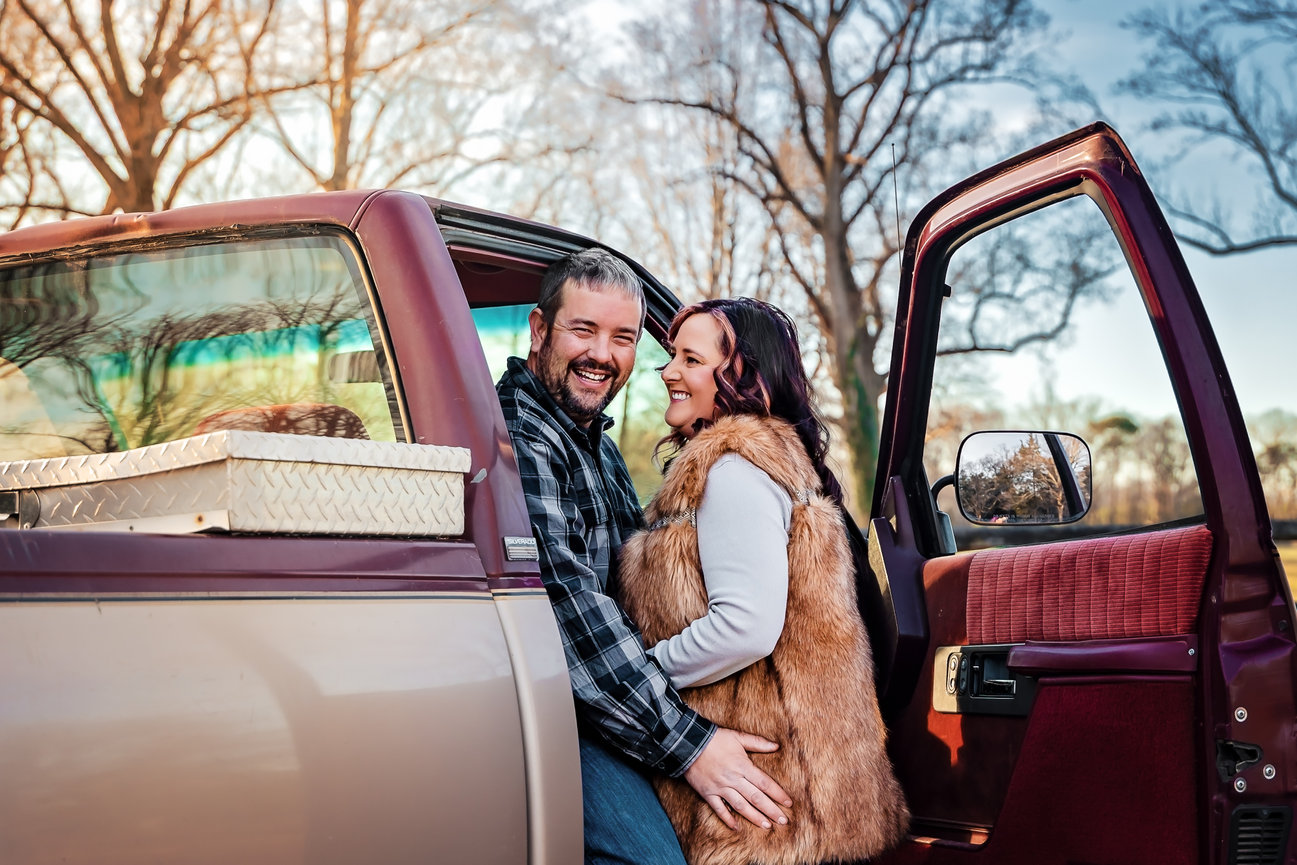 An outdoor couple photography session of a couple smiling in an open truck door, surrounded by bare trees.
