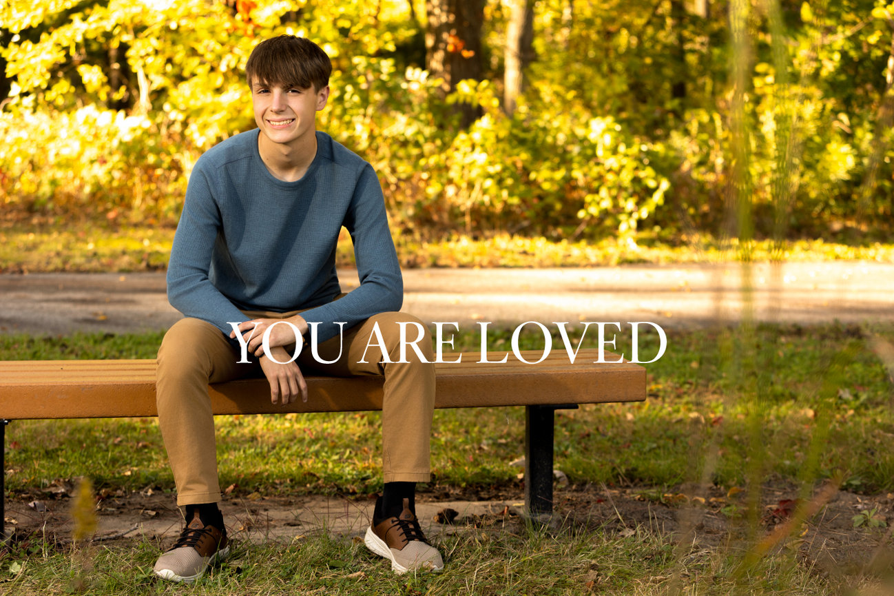 Smiling person sitting on a bench in a park with autumn trees, text reads YOU ARE LOVED.