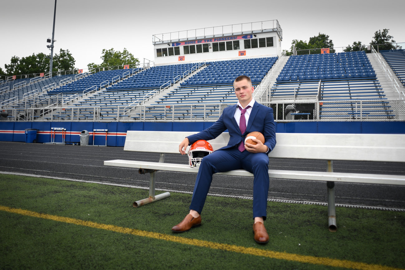 A young man in a suit sits on a bench at a football stadium, holding a football and wearing a helmet nearby.
