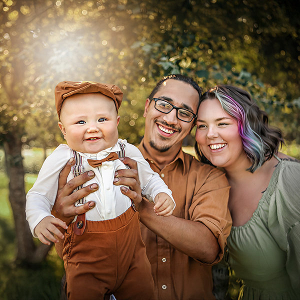 Smiling family with baby in nature, sunlight filtering through trees.