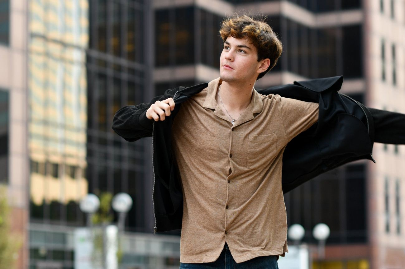 Teen boy stands confidently outdoors in a cityscape, wearing a black jacket and brown shirt.