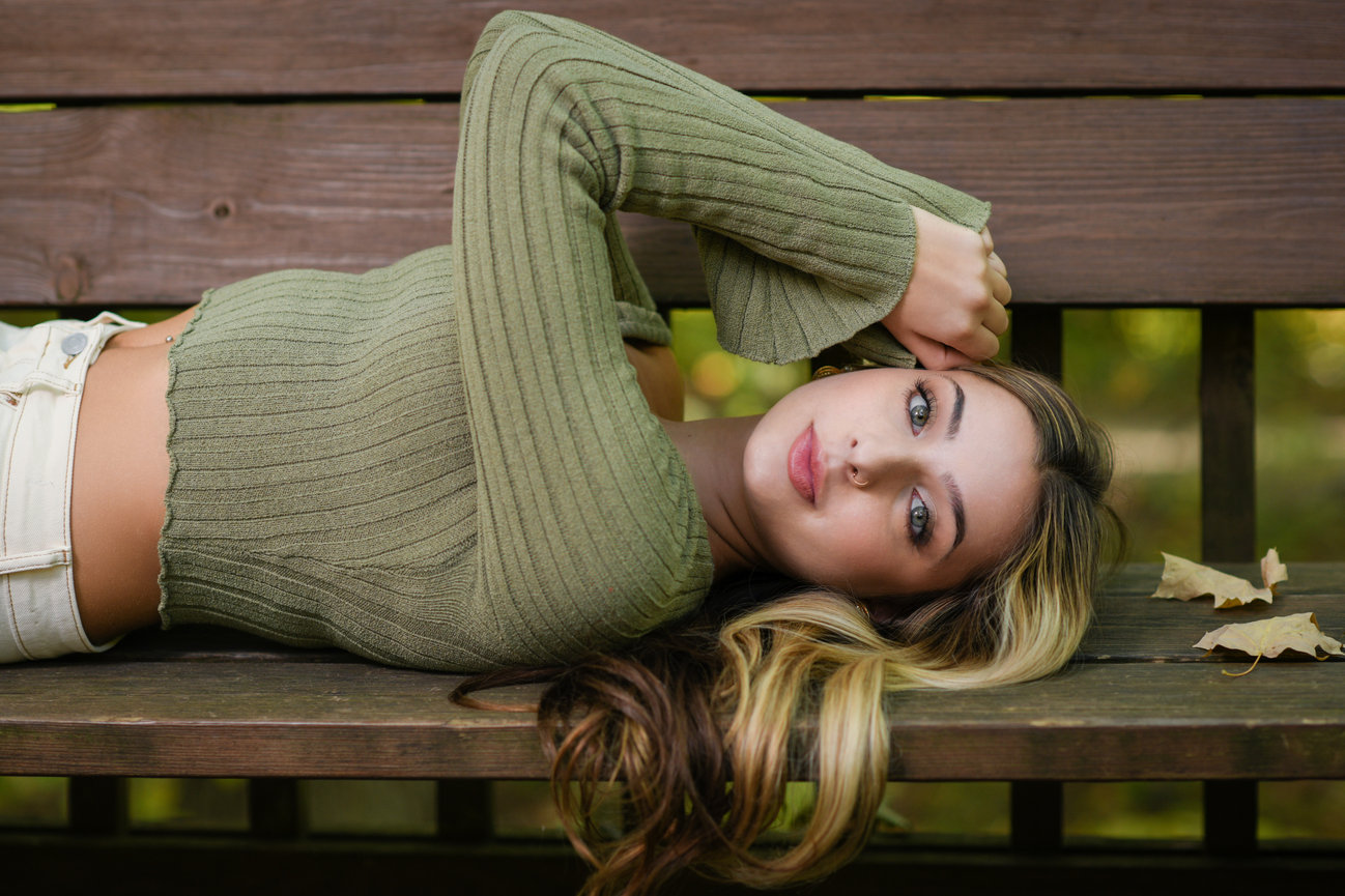 A teenage girl with long blonde hair and a green sweater laying on a bench with fall leaves around her for senior pictures in Columbus OH.